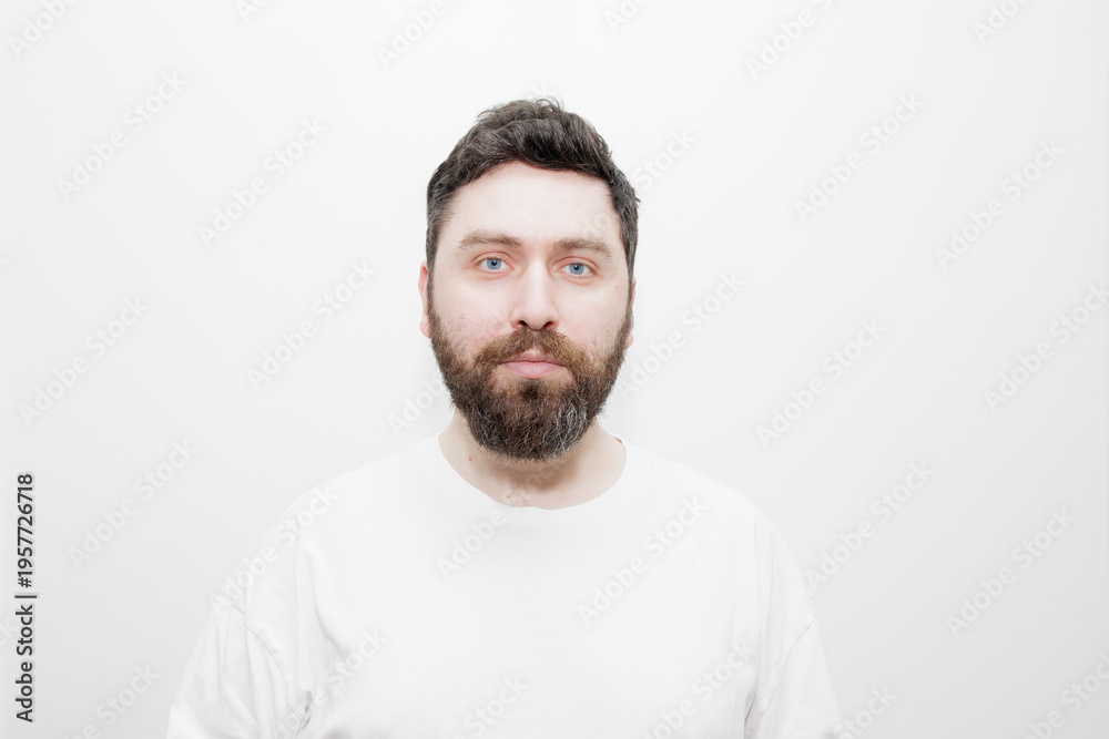 Obraz premium A direct, neutral headshot portrait of a man with dark hair, beard and blue eyes in a white t-shirt on a white background. Stock photo