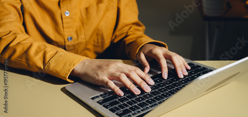 Person typing on laptop keyboard wearing orange shirt, close up of hands working on computer, indoor workspace with focused activity and warm lighting