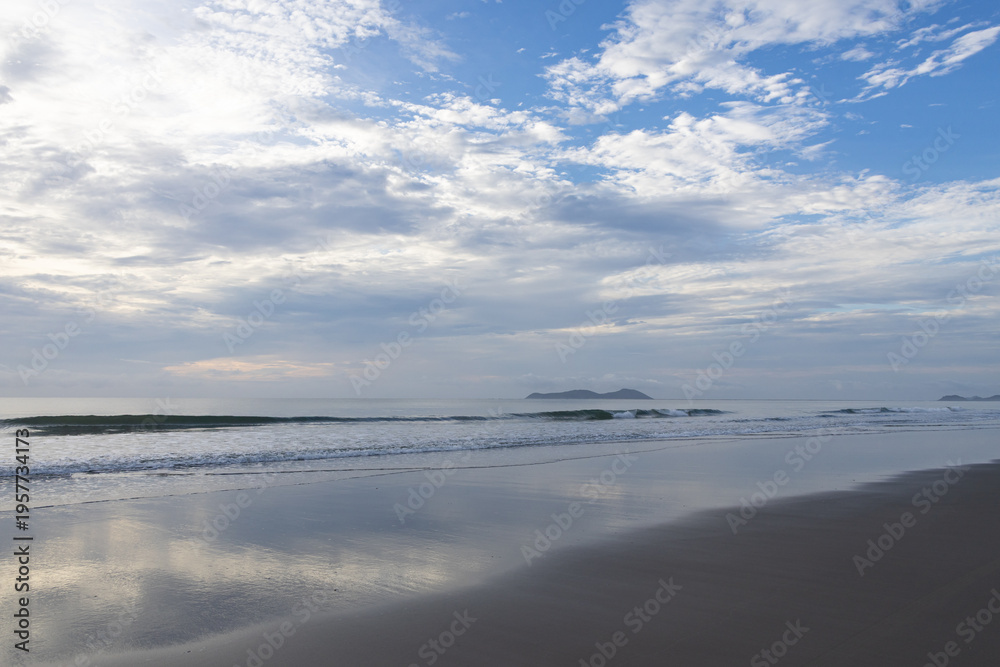 Fototapeta premium Serene coastal landscape with gentle waves on wet, reflective sand under a soft, cloudy sky. 