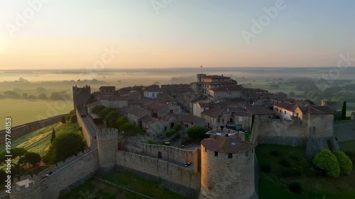 An aerial view of a walled town at dawn. Fog blankets the fields, and the sun shines