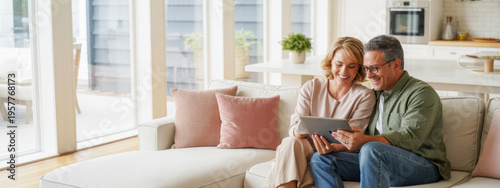 Candid photo of happy middle aged couple using digital tablet relaxing on couch at home. Smiling mature man and woman holding tab browsing internet on pad device sitting on sofa in living room. 