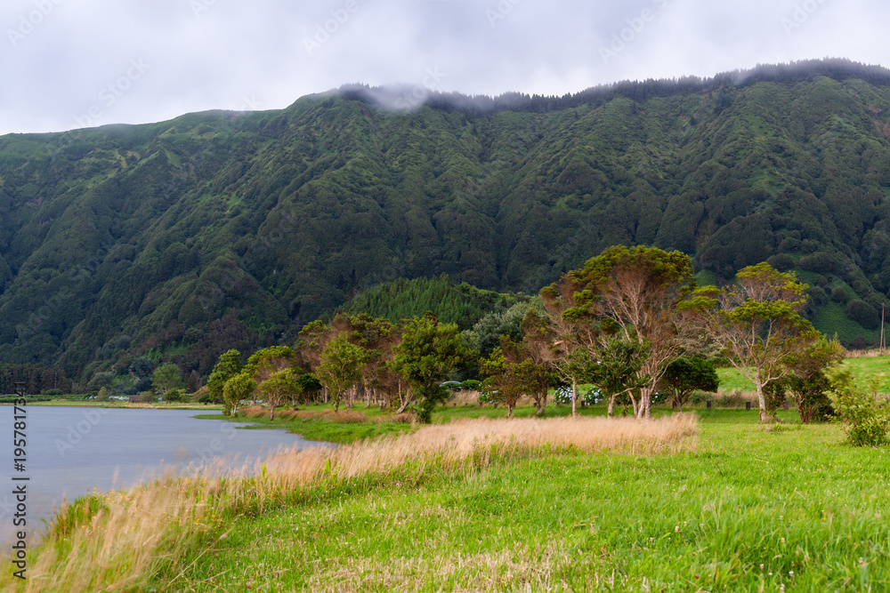Fototapeta premium Lush green hills of Sete Cidades, San Miguel Island, Azores, Portugal. Calm lake surrounded by forested mountains. Cloudy sky over serene landscape.