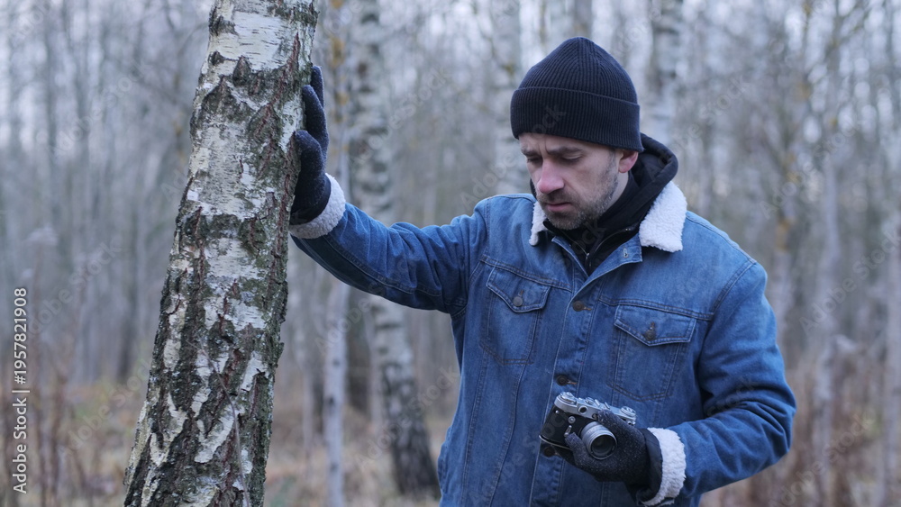 Obraz premium Man holding a vintage camera, dressed in winter clothing including a beanie, denim jacket, and gloves, looking down while standing next to a birch tree trunk in a sparse winter forest