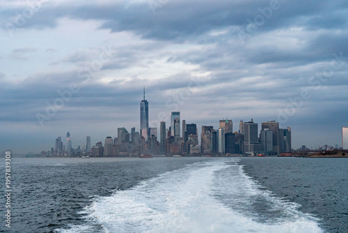 Lower Manhattan skyline of New York City as seen from New York Harbor. Iconic skyscrapers, including One World Trade Center, rise against a backdrop of cloudy skies