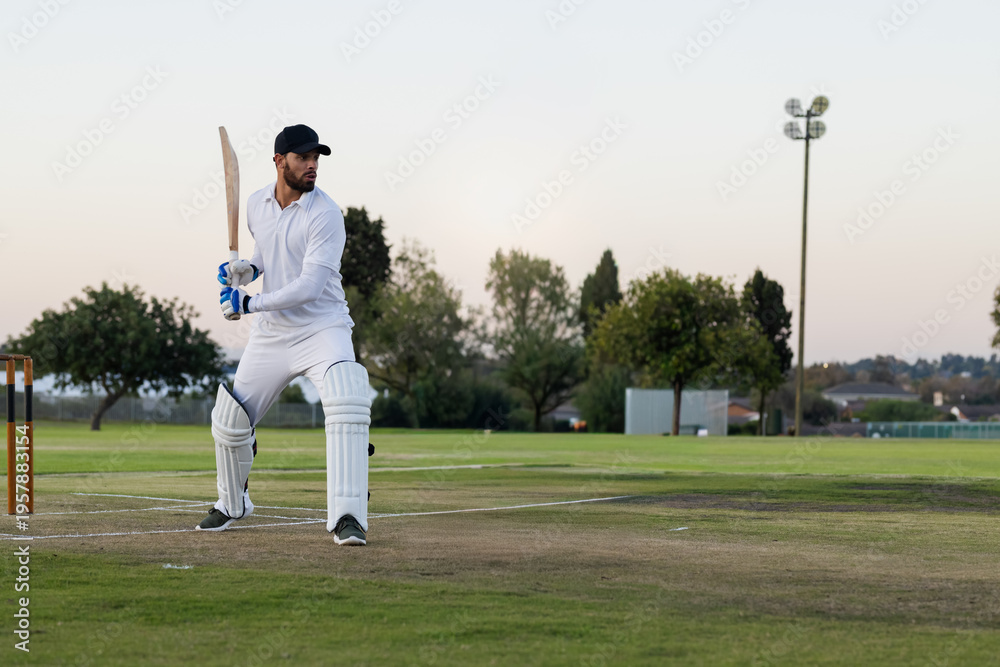Obraz premium Male batsman in white kit standing at crease near stumps at dusk holding bat, copy space