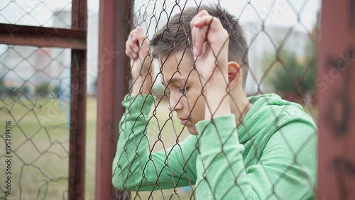 Fotografie Teenage Boy Holding Fence with Sad Expression Outdoors, have fun and enjoy