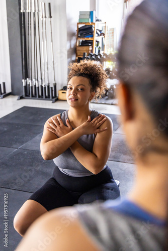 Two adult females training in athletic wear near barbell rack, one kneeling crossing arms on mats