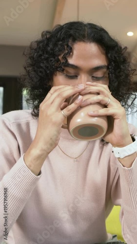 Vertical video: Reaching nonbinary adult in pink sweater grabbing mug on couch, sipping, relaxing