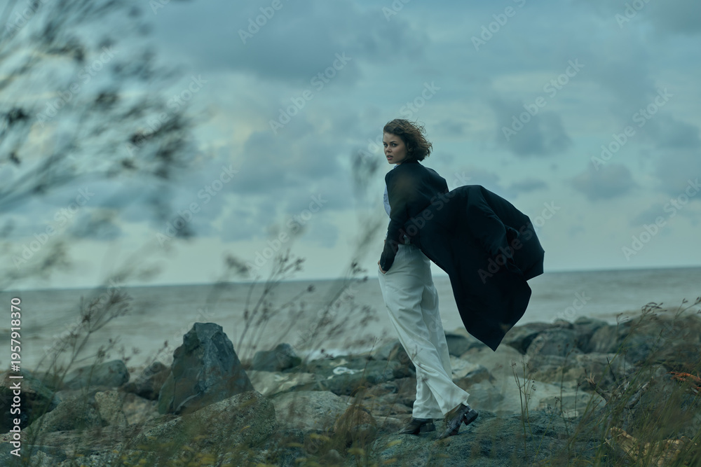 Fototapeta premium windy coastline scene with a woman walking along rocky shore, flowing coat catching the breeze, ocean horizon in the distance, moody blue tones and natural outdoor atmosphere