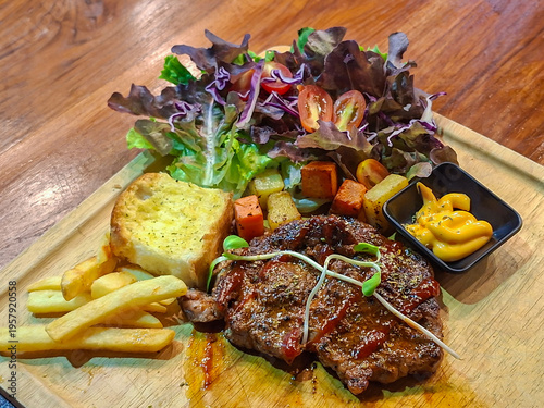 A steak arranged on a wooden cutting board, served with leafy green salad, toasted garlic bread, french fries, and an appetizing yellow sauce in a black cup, resembling cheese or mustard.