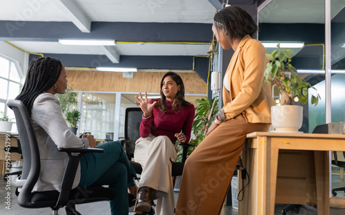 Diverse female coworkers discussing project ideas at open-plan office corner with coffee mug