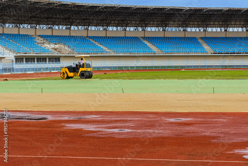 Road roller vehicle working and compacting soil and freshly installed natural sod on new sports field inside large stadium. Professional lawn installation in progress at stadium construction site.