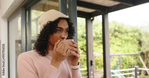 Non-binary adult sipping pale mug while spotting garden through sliding door by balcony, copy space