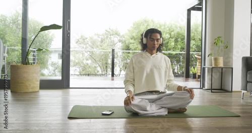 Non-binary person tapping smartphone, donning headphones and meditating on green mat in living room