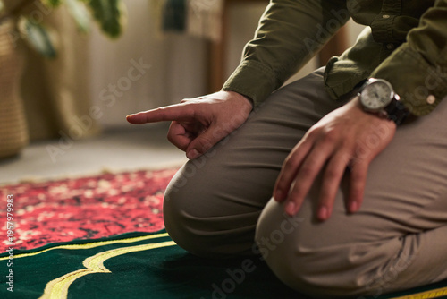 Fotografie Young Muslim man kneeling on prayer rug raising finger during Islamic prayer rit