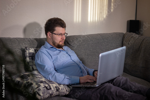 Man working on laptop sitting on couch in cozy home environment casually completing tasks after workday in comfortable home atmosphere