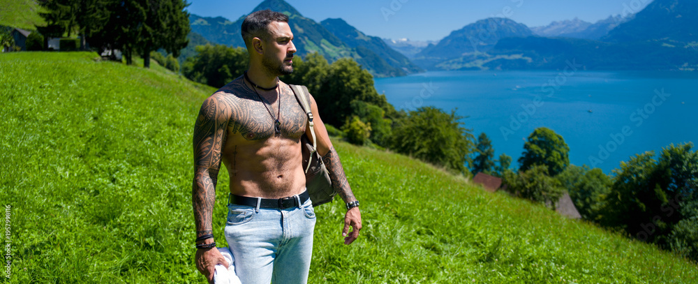 Fototapeta premium Travel portrait with muscular man hiking mountain path. Travel adventure with confident man near alpine lake. Travel lifestyle with active hiker walking meadow. Travel inspiration in summer nature.