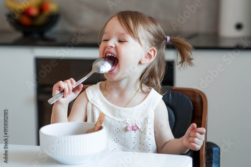 A two-year-old girl is sitting at the table eating porridge or soup from a white plate. Baby food. Feeding a child. Healthy food for children.