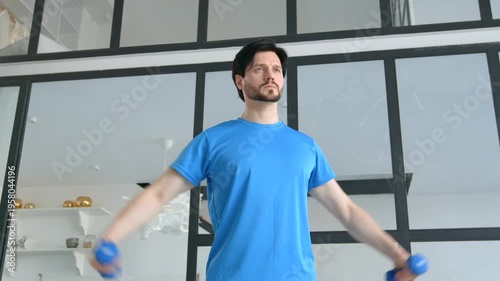 Motivated healthy lifestyle fitness man in sportswear practicing dumbbell lateral raise exercise, strengthening and toning shoulder and upper body muscles in domestic room.