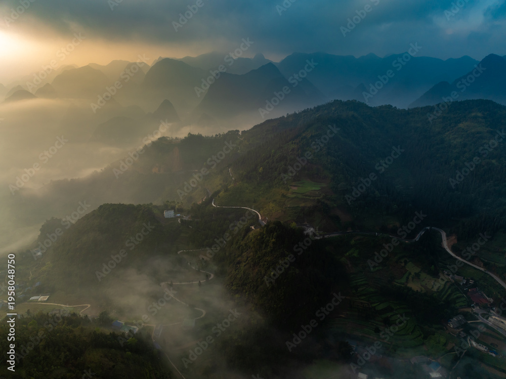 Fototapeta premium An aerial view shows the winding roads and misty mountains of Dong Van, Ha Giang, Vietnam, as the sun rises, creating a scenic and ethereal landscape at dawn.
