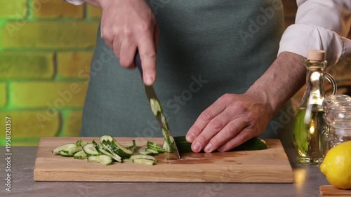 A male chef slices a cucumber with a knife on a cutting board to prepare a salad.