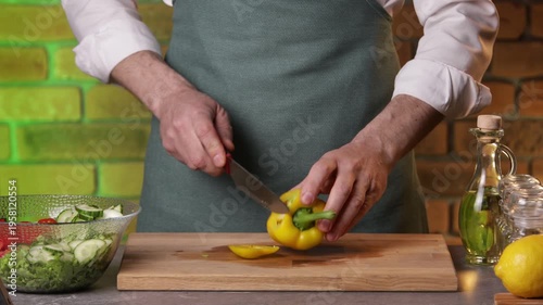 A male chef slices yellow bell pepper with a knife on a cutting board to prepare a salad.