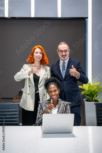 Diverse business team showing thumbs up with seated woman using laptop and standing managers in modern office