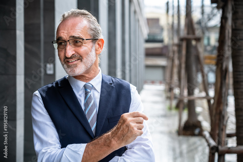 Smiling mature Caucasian businessman pointing sideways with hand gesture in modern city wearing formal suit and glasses
