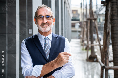 Confident mature Caucasian businessman smiling with crossed arms and pointing gesture in modern city wearing formal suit and glasses, looking at camera