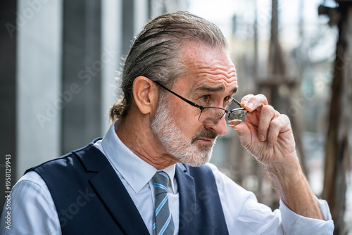 Serious mature Caucasian businessman adjusting glasses with hand focused sideways posture wearing formal suit outdoors in modern city