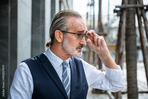 Confident mature Caucasian businessman adjusting glasses with hand thoughtful sideways posture wearing formal suit outdoors in modern city
