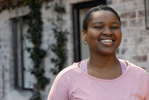 Female standing smiling wearing pink top with lavender straps and gold hoops by brick facade