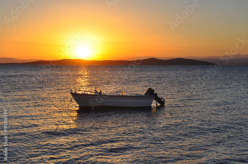 Small motorboat anchored on calm sea during golden sunset with warm light reflecting on the water and distant island silhouettes on the horizon