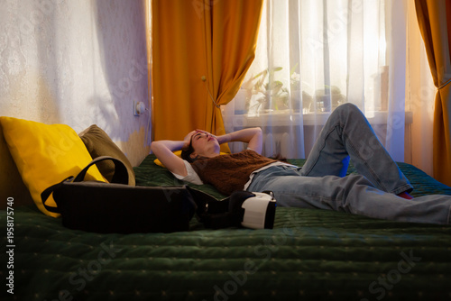 Young woman with long hair lies on bed and holds VR device with irritated expression. Futuristic technology in real life and the emotional impact of digital use. Mid shot