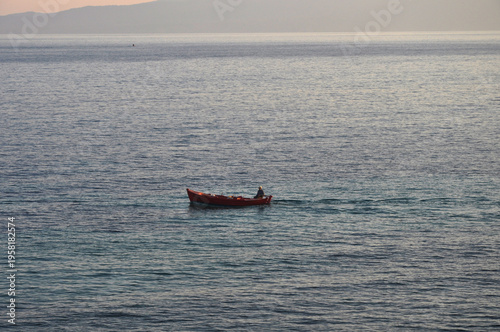 A solitary fisherman in a small red wooden boat floating on a calm sea with soft light, distant coastline and quiet Mediterranean atmosphere