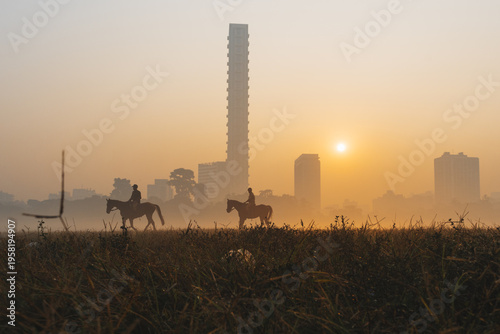 View of silhouetted riders astride horses traversing a sun-kissed field, with the iconic, towering 42 building piercing through the misty Kolkata skyline, Kolkata, West Bengal, India.