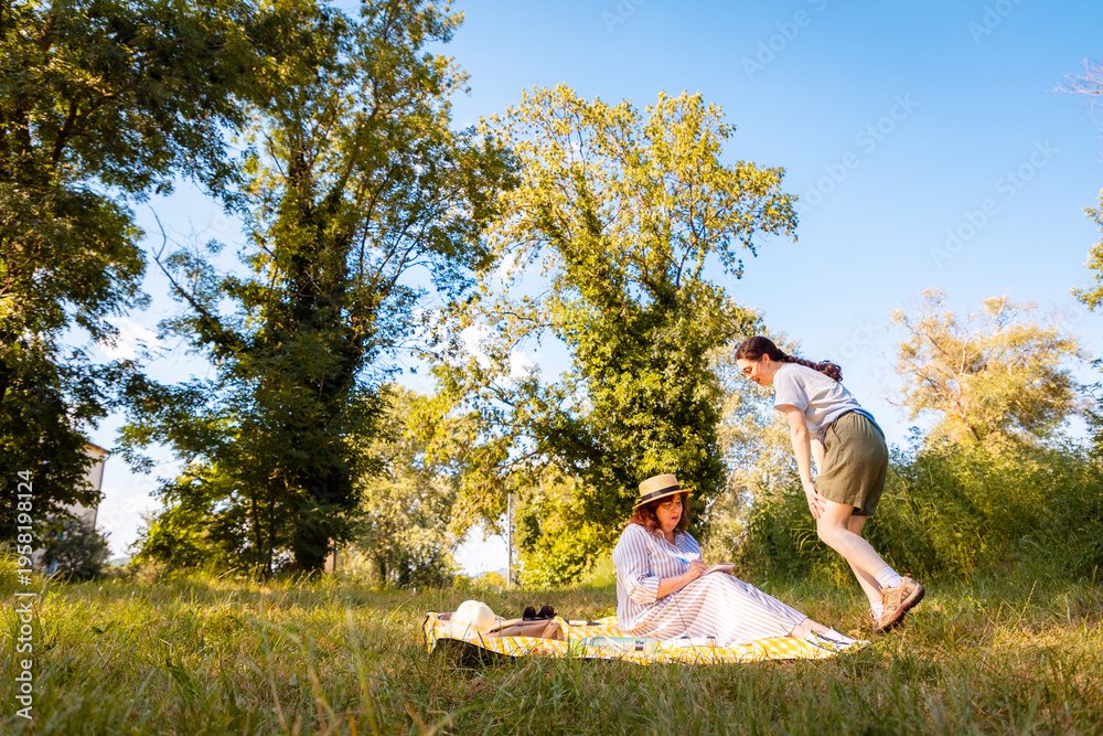 © _KUBE_ - Outdoor relaxation with mother and daughter on vacation, bonding on a yellow blanket under the trees. Wide shot, low angle view