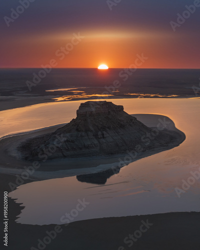 Aerial view of a solitary rock formation rising from shallow waters under a vibrant sunset, Aktau, Mangystau Region, Kazakhstan.