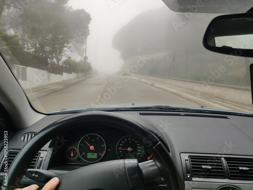 Driver's point of view from inside a car cockpit driving through a residential street with heavy fog and low visibility during the morning.