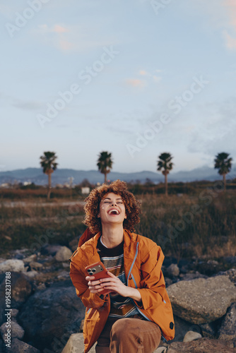 Happy woman with curly hair wearing orange jacket laughs holding smartphone outdoors near rocks with palm trees and mountains in background during casual lifestyle moment.