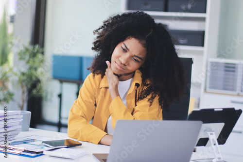 Businesswoman experiencing discomfort and pain in her lower back while sitting at her desk, working on a laptop in a modern office environment, highlighting work-related health issues and stress