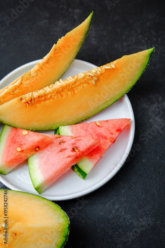 Tablou pe pânză Melon slices and watermelon pieces arranged on a white plate against a dark back