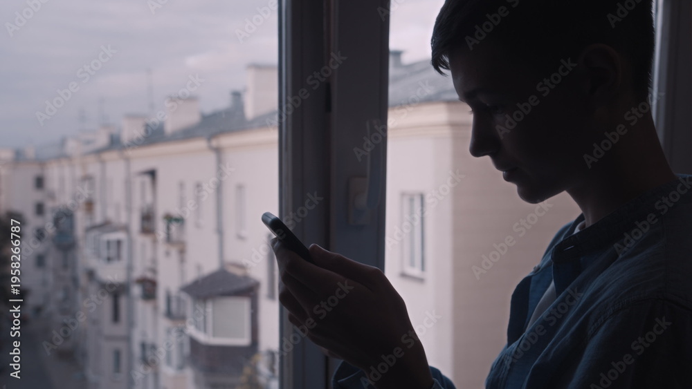 Fototapeta premium Young Man Using Smartphone Indoors by Window at Dusk, have fun and enjoy