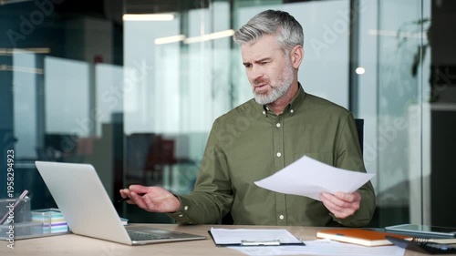 Confused mature businessman sitting at desk in office, holding document and staring at laptop screen with puzzled expression, struggling to understand financial report and data, uncertainty and stress