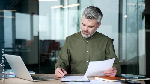 Mature gray-haired businessman sitting at desk in modern office, holding document in hand and writing on clipboard, filling business form, taking notes, focused professional managing office paperwork.