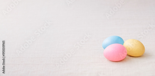 Three colored Easter eggs in blue, pink, and yellow lying on a light beige linen fabric with copy space. Minimalist composition for spring holiday, celebration, and tradition