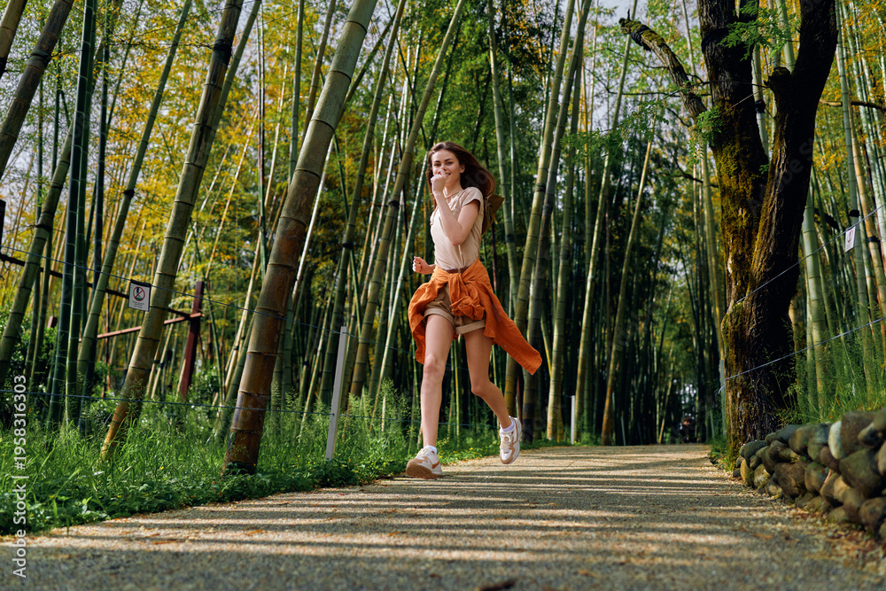 Naklejka premium Woman running along a bamboo forest path, smiling and jogging with smartphone and headphones. Casual outfit with shorts and sweater tied around waist during outdoor fitness session.