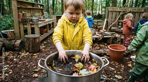 Child playing in mud kitchen with bare hands dipping into a pot of wet soil and autumn leaves during outdoor forest school activity.