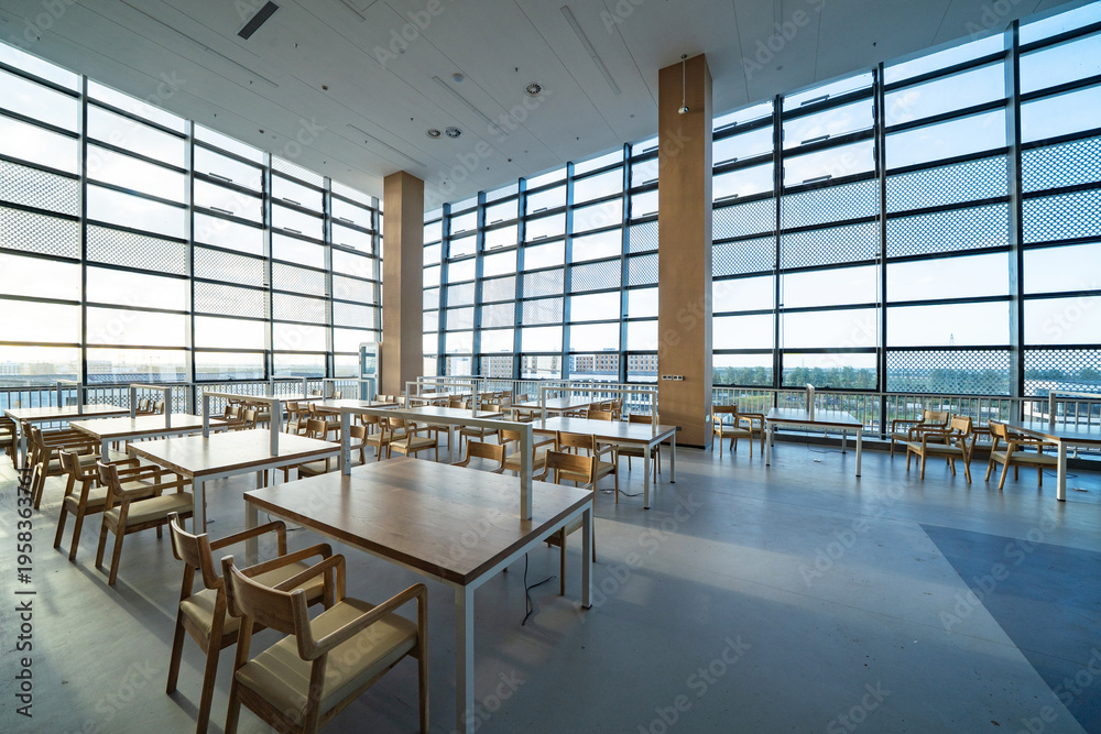 Fototapeta premium Modern library reading room with empty wooden tables and chairs under large glass curtain walls