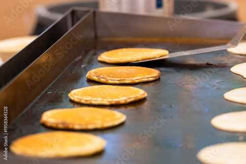 Canvastavla A neat row of pancakes fills a flat top grill, and at the far end a long spatula is mid-flip, catching the furthest pancake in the moment of rotation